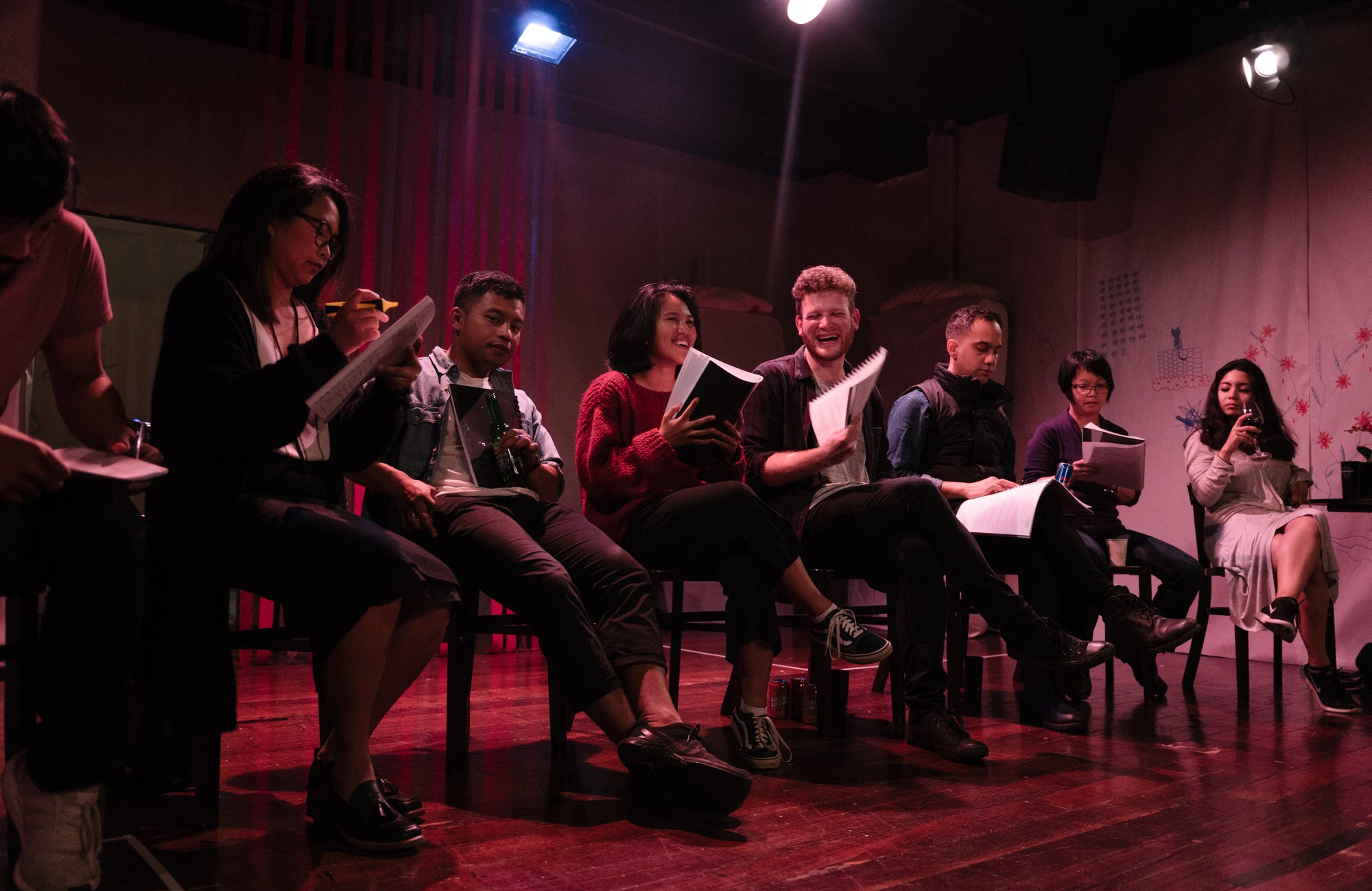 A group of people sitting in a line on stools under warm lights. They are holding scripts and smiling.